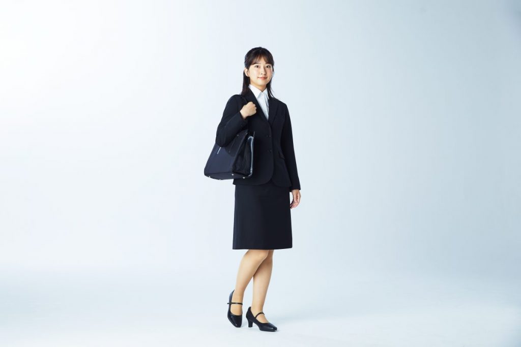 リクルートスーツスタイルの女性が、右肩にバッグをかけて、こちらを向いて立っている。