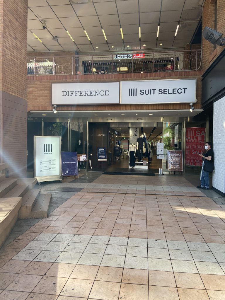 スーツセレクト横浜西の店頭画像。
店頭正面の画像。
ガラス張り、正面にマネキン人形2～3体が見える。difference、suit　select両店の看板が入口上に掲げられている。看板はいずれも白地に黒。向かって左側にセレクトのつい立て看板あり。全体的に煉瓦調。

