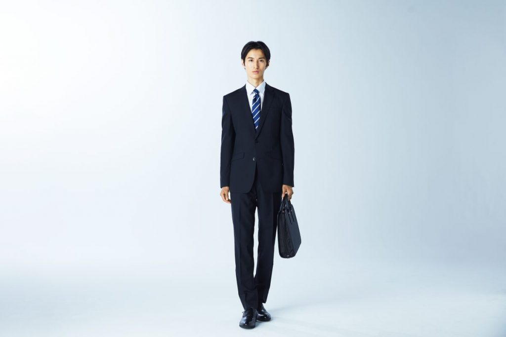 リクルートスーツスタイルの男性が、こちらを向いて立っている。ネイビー系ストライプ柄ネクタイ着用。左手にはバッグ。